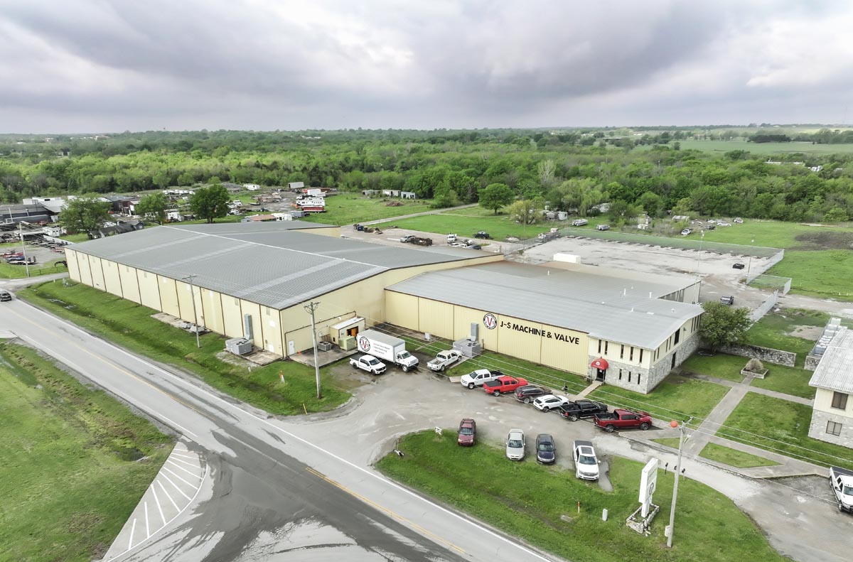 Aerial view of a large industrial warehouse with the sign S&S Machine & Valve on the front. Several cars and trucks are parked outside, surrounded by greenery and a partly cloudy sky.
