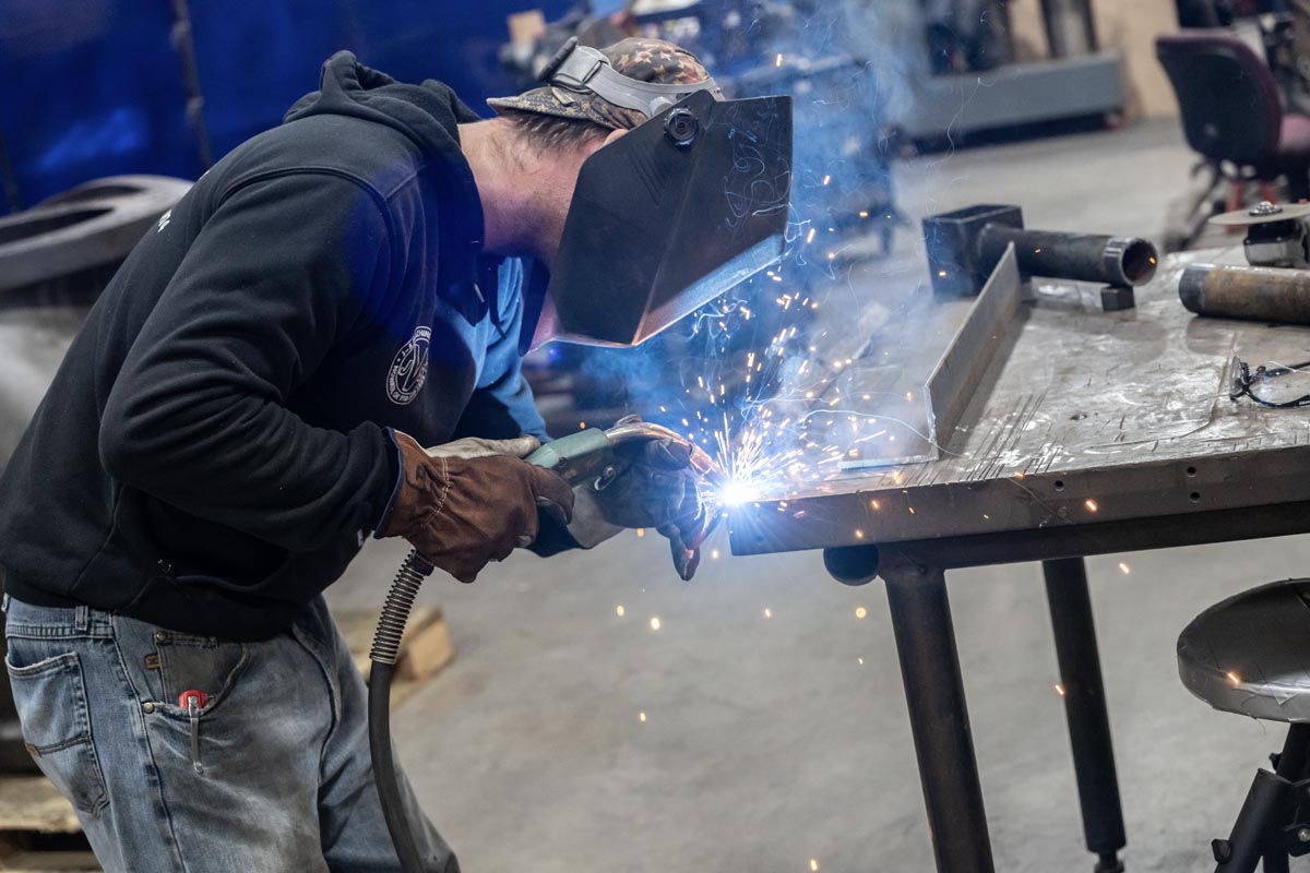 A person wearing protective gear uses a welding tool on a metal table, producing bright sparks in a workshop setting.