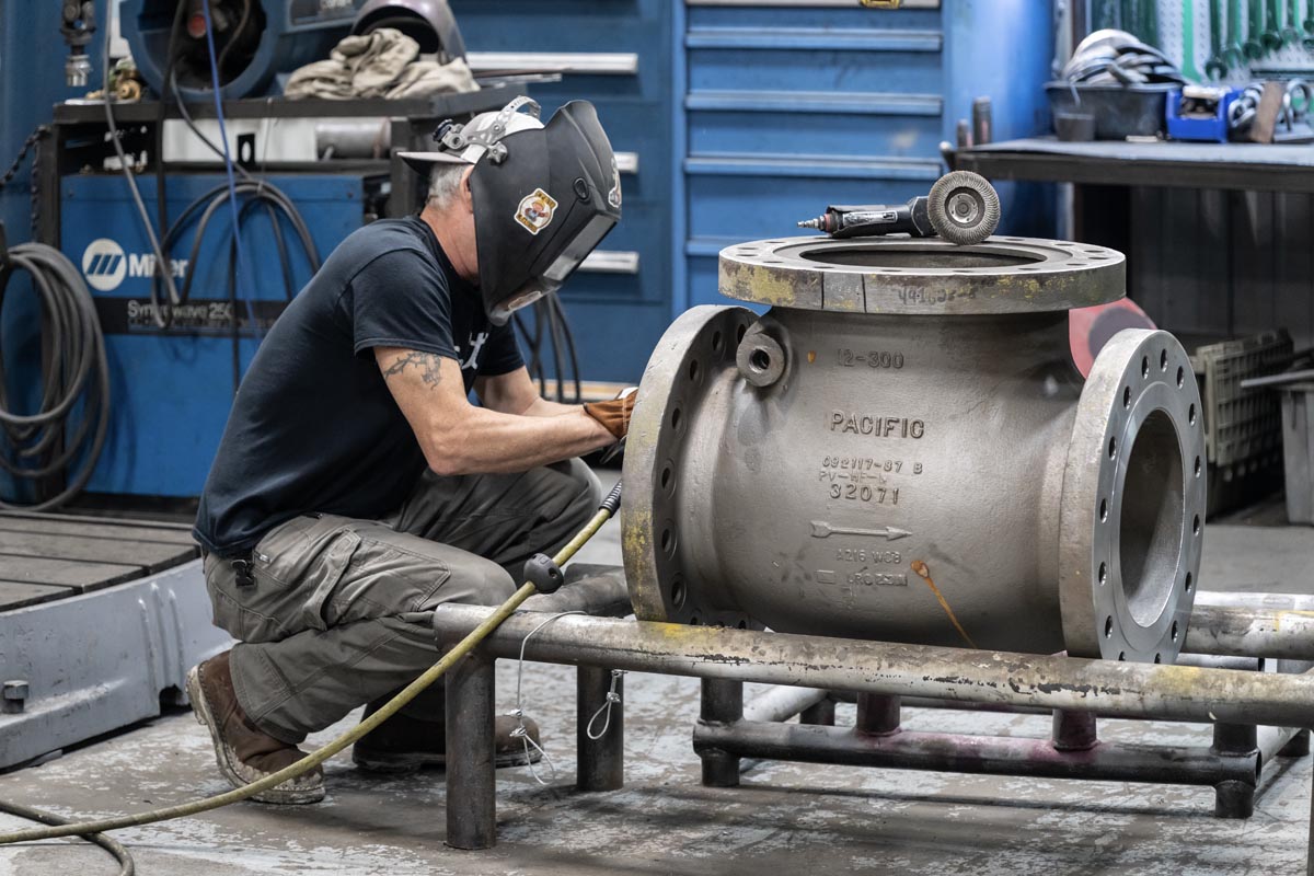 A worker wearing a welding helmet and gloves welds a large industrial valve in a workshop, surrounded by tools and equipment.