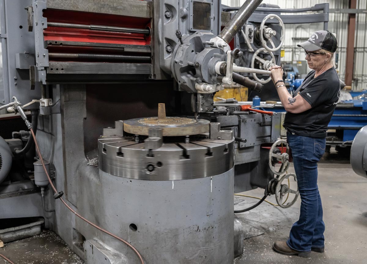 A person wearing glasses and a cap operates a large industrial machine in a factory setting, adjusting controls while standing beside the spinning metal workpiece.
