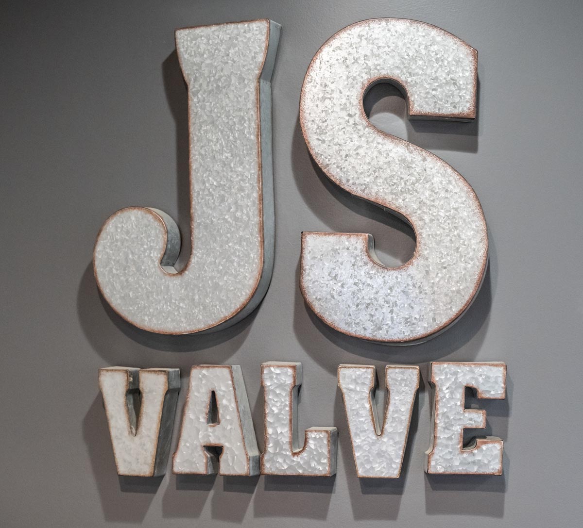 Metallic block letters mounted on a gray wall spell out JS VALVE, with JS in large letters above VALVE in smaller size. The letters have a textured, galvanized appearance with a rustic finish.