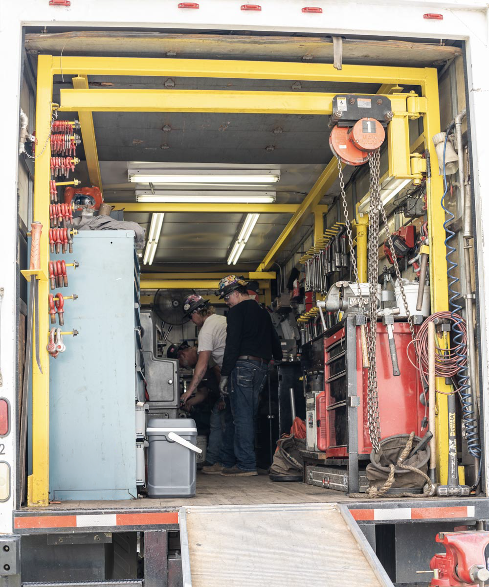 Two workers stand inside a mobile workshop truck filled with tools, equipment, and machinery. The interior has shelves, hooks, and organized red toolboxes along both sides under fluorescent lighting.