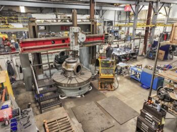 Large industrial machine shop with various equipment, workstations, tools, and machinery. A worker operates a massive vertical lathe with a large metal part. The space is organized with metal shelves and safety barriers. Large industrial machine shop with various equipment, workstations, tools, and machinery. A worker operates a massive vertical lathe with a large metal part. The space is organized with metal shelves and safety barriers.