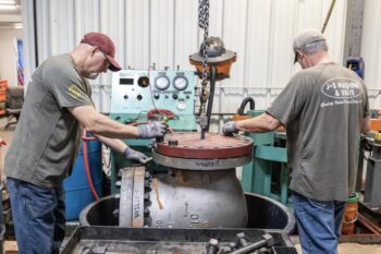 Two workers in a workshop use a chain hoist to lift the heavy lid of a large industrial valve. They wear gloves and T-shirts with “J-S Machine & Valve” on the back. Industrial equipment and tools are visible around them. Two workers in a workshop use a chain hoist to lift the heavy lid of a large industrial valve. They wear gloves and T-shirts with “J-S Machine & Valve” on the back. Industrial equipment and tools are visible around them.