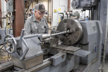An older man wearing glasses and a cap operates a large industrial lathe machine in a workshop, shaping a metal rod. The environment is filled with machinery and workstations in the background. An older man wearing glasses and a cap operates a large industrial lathe machine in a workshop, shaping a metal rod. The environment is filled with machinery and workstations in the background.