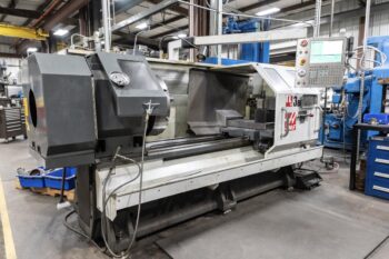 A large industrial CNC lathe machine is situated in a workshop, surrounded by tools and equipment, with a control panel on the right and metal parts visible on the machine bed. A large industrial CNC lathe machine is situated in a workshop, surrounded by tools and equipment, with a control panel on the right and metal parts visible on the machine bed.