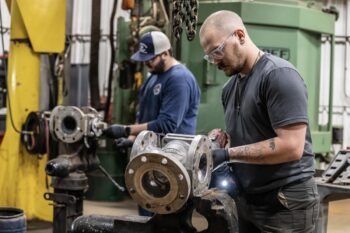 Two men work on large metal machine parts in an industrial workshop. One man in the foreground wears safety glasses and gloves while using a tool; another man works in the background. Machinery and equipment surround them. Two men work on large metal machine parts in an industrial workshop. One man in the foreground wears safety glasses and gloves while using a tool; another man works in the background. Machinery and equipment surround them.