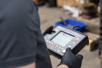 A person wearing black gloves holds an electronic control device with a display screen and multiple colorful buttons, likely operating machinery in an industrial setting. A person wearing black gloves holds an electronic control device with a display screen and multiple colorful buttons, likely operating machinery in an industrial setting.