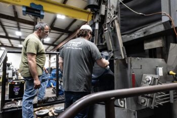 Two men work together in a factory, operating a large industrial machine. One stands on a step, observing closely, while the other adjusts controls. The setting is busy, with equipment and tools visible around them. Two men work together in a factory, operating a large industrial machine. One stands on a step, observing closely, while the other adjusts controls. The setting is busy, with equipment and tools visible around them.