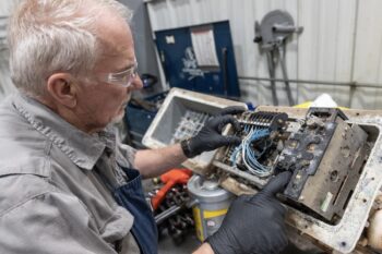 An older man wearing safety glasses and black gloves repairs a dusty, open electrical control panel with exposed wires in a workshop. He uses one hand to point at the components inside the panel. An older man wearing safety glasses and black gloves repairs a dusty, open electrical control panel with exposed wires in a workshop. He uses one hand to point at the components inside the panel.