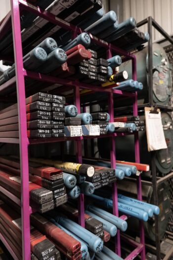 Metal rods with blue and red ends are stored horizontally on a purple rack, each labeled with white stickers and black writing, inside an industrial workshop. Manufacturing equipment is visible in the background. Metal rods with blue and red ends are stored horizontally on a purple rack, each labeled with white stickers and black writing, inside an industrial workshop. Manufacturing equipment is visible in the background.