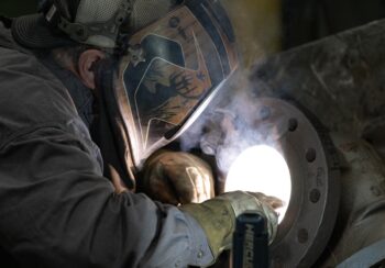 A welder wearing a protective helmet and gloves is working on a metal pipe, with bright sparks and smoke rising from the welding process. A welder wearing a protective helmet and gloves is working on a metal pipe, with bright sparks and smoke rising from the welding process.