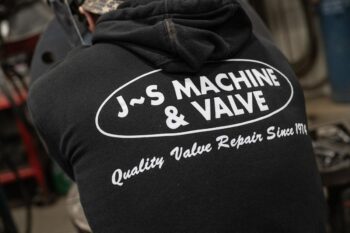 A person wearing a black hoodie with the text J~S MACHINE & VALVE, Quality Valve Repair Since 1991 printed in white on the back. Workshop equipment is blurred in the background. A person wearing a black hoodie with the text J~S MACHINE & VALVE, Quality Valve Repair Since 1991 printed in white on the back. Workshop equipment is blurred in the background.