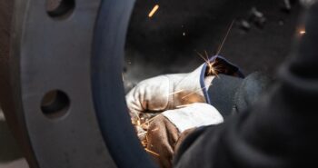 A close-up of a person wearing protective gloves using a tool to grind or cut metal, creating bright orange sparks. The image is framed through a circular metal structure. A close-up of a person wearing protective gloves using a tool to grind or cut metal, creating bright orange sparks. The image is framed through a circular metal structure.