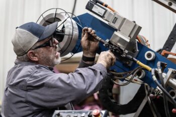 A man wearing glasses, a cap, and work clothes operates and adjusts a large industrial machine with wires and metal components inside a workshop. A man wearing glasses, a cap, and work clothes operates and adjusts a large industrial machine with wires and metal components inside a workshop.