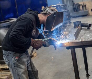 A person wearing a welding mask and gloves is using a welding torch on a metal table, with bright sparks flying in a workshop setting. A person wearing a welding mask and gloves is using a welding torch on a metal table, with bright sparks flying in a workshop setting.
