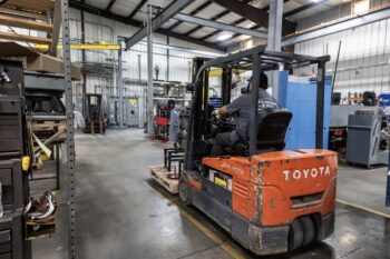 A person operates an orange Toyota forklift inside a spacious industrial warehouse with shelves, machinery, and equipment visible in the background. A person operates an orange Toyota forklift inside a spacious industrial warehouse with shelves, machinery, and equipment visible in the background.
