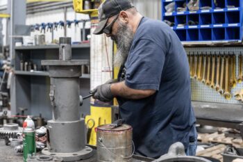 A man with a beard and glasses, wearing a cap and gloves, works on a large metal component in an industrial workshop. Tools and equipment are organized on the wall and workbench around him. A man with a beard and glasses, wearing a cap and gloves, works on a large metal component in an industrial workshop. Tools and equipment are organized on the wall and workbench around him.
