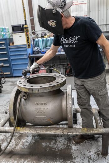 A man wearing a welding helmet and a black Peterbilt t-shirt inspects a large industrial valve in a workshop, using a tool while standing next to blue cabinets and equipment. A man wearing a welding helmet and a black Peterbilt t-shirt inspects a large industrial valve in a workshop, using a tool while standing next to blue cabinets and equipment.