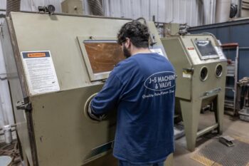 A man wearing a blue J-S Machine & Valve shirt operates a sandblasting machine inside an industrial workshop, standing in front of a large metal cabinet with warning labels. A man wearing a blue J-S Machine & Valve shirt operates a sandblasting machine inside an industrial workshop, standing in front of a large metal cabinet with warning labels.