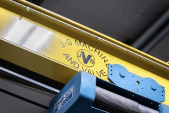 A close-up of a yellow overhead crane beam with the text J S MACHIN AND VALVE and a circular JVS logo in black. Below is a blue industrial hoist marked SHAW-BOX inside a warehouse. A close-up of a yellow overhead crane beam with the text J S MACHIN AND VALVE and a circular JVS logo in black. Below is a blue industrial hoist marked SHAW-BOX inside a warehouse.
