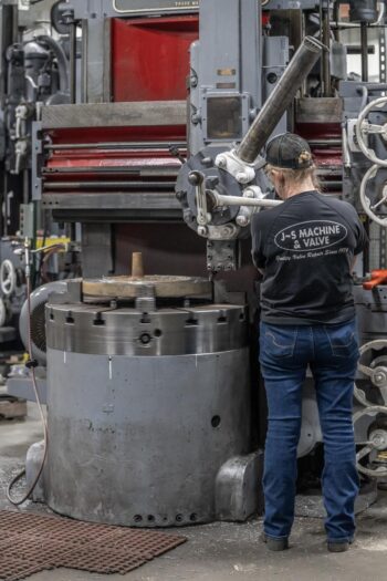 A person wearing a J-S Machine & Valve shirt operates a large industrial machine in a workshop, adjusting controls on the equipment. The setting is busy with metal machinery and tools visible in the background. A person wearing a J-S Machine & Valve shirt operates a large industrial machine in a workshop, adjusting controls on the equipment. The setting is busy with metal machinery and tools visible in the background.