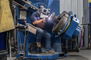 A worker wearing a protective helmet uses welding equipment to join metal parts on a large rotating machine inside an industrial workshop. A worker wearing a protective helmet uses welding equipment to join metal parts on a large rotating machine inside an industrial workshop.