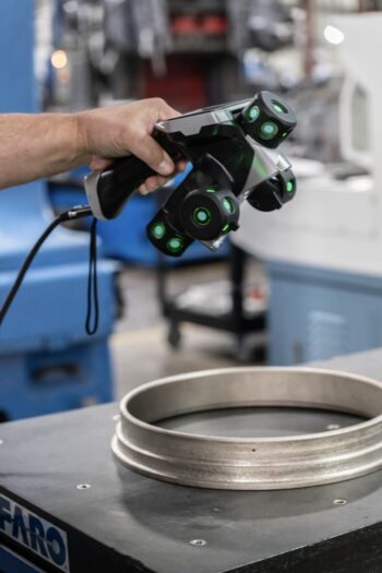 A person uses a handheld 3D scanner with glowing green lights to scan a circular metal component in an industrial setting. A person uses a handheld 3D scanner with glowing green lights to scan a circular metal component in an industrial setting.