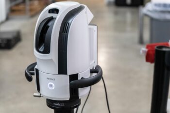 A close-up of a white and black KEYENCE industrial device, possibly a scanner or sensor, mounted on a stand in a large indoor workspace with a concrete floor. A close-up of a white and black KEYENCE industrial device, possibly a scanner or sensor, mounted on a stand in a large indoor workspace with a concrete floor.