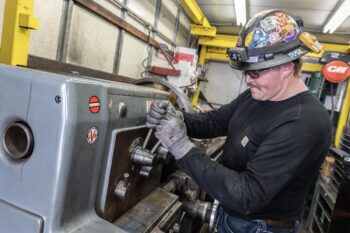A worker in a black shirt, safety glasses, gloves, and a helmet with stickers uses a wrench on industrial machinery inside a workshop. Yellow metal beams and various equipment are visible in the background. A worker in a black shirt, safety glasses, gloves, and a helmet with stickers uses a wrench on industrial machinery inside a workshop. Yellow metal beams and various equipment are visible in the background.