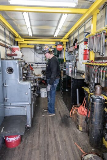 A person wearing safety glasses and a hard hat operates machinery inside a narrow, organized workshop filled with tools, equipment, and storage shelves. A person wearing safety glasses and a hard hat operates machinery inside a narrow, organized workshop filled with tools, equipment, and storage shelves.